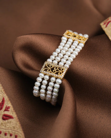 The Divine Pearl Shine Bracelet by Chandrani Pearls, a multi-strand white pearl piece with gold rectangular clasps, is elegantly displayed on brown satin fabric with embroidered trim.