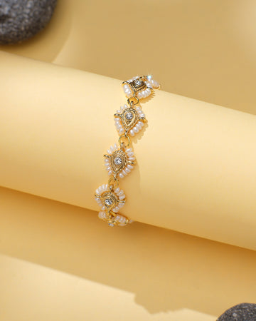 The Dainty Pearl Bracelet with CZ Halo Accents by Chandrani Pearls features teardrop-shaped charms, white pearl-like beads, and sparkling cubic zirconia, elegantly displayed on a rolled yellow surface.
