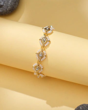 The Dainty Pearl Bracelet with CZ Halo Accents by Chandrani Pearls features teardrop-shaped charms, white pearl-like beads, and sparkling cubic zirconia, elegantly displayed on a rolled yellow surface.