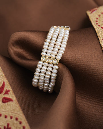 The Classic Elegance Bracelet by Chandrani Pearls features luminous multi-strand pearls and a gold, diamond-accented clasp, elegantly displayed on brown satin.