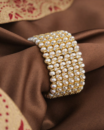 The Celestial Light CZ Pearl Bracelet by Chandrani Pearls features rows of white pearls and sparkling CZ stones on wide gold, shown on brown fabric—a celestial-inspired statement piece.