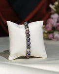 The Elegant Black 1 line Bracelet by Chandrani Pearls India features dark, irregular pearls with subtle silver accents, exuding sophistication as it rests on a white cushion with softly blurred floral decor in the background.