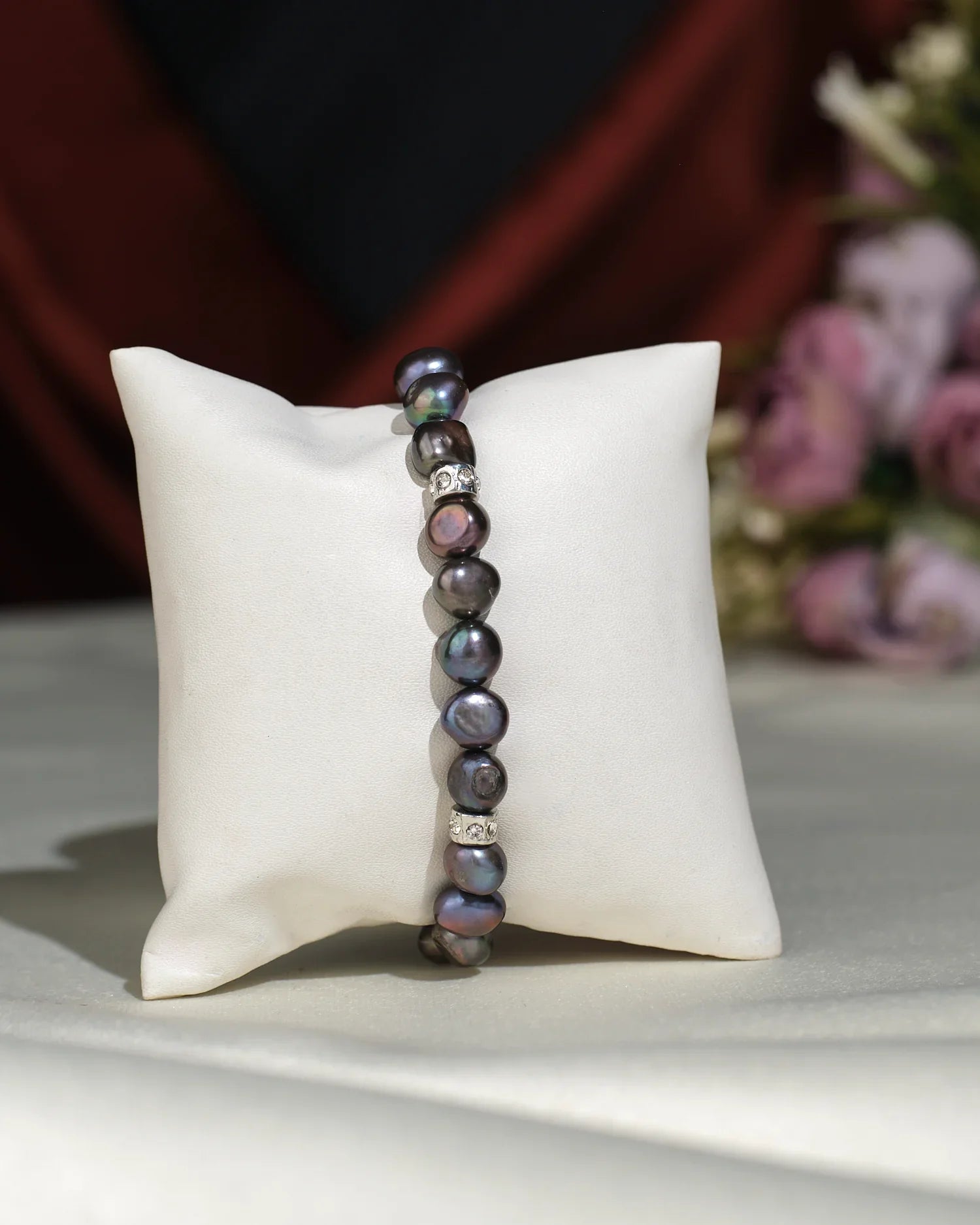 The Elegant Black 1 line Bracelet by Chandrani Pearls India features dark, irregular pearls with subtle silver accents, exuding sophistication as it rests on a white cushion with softly blurred floral decor in the background.