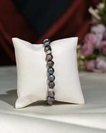 The Elegant Black 1 line Bracelet by Chandrani Pearls India features dark, irregular pearls with subtle silver accents, exuding sophistication as it rests on a white cushion with softly blurred floral decor in the background.