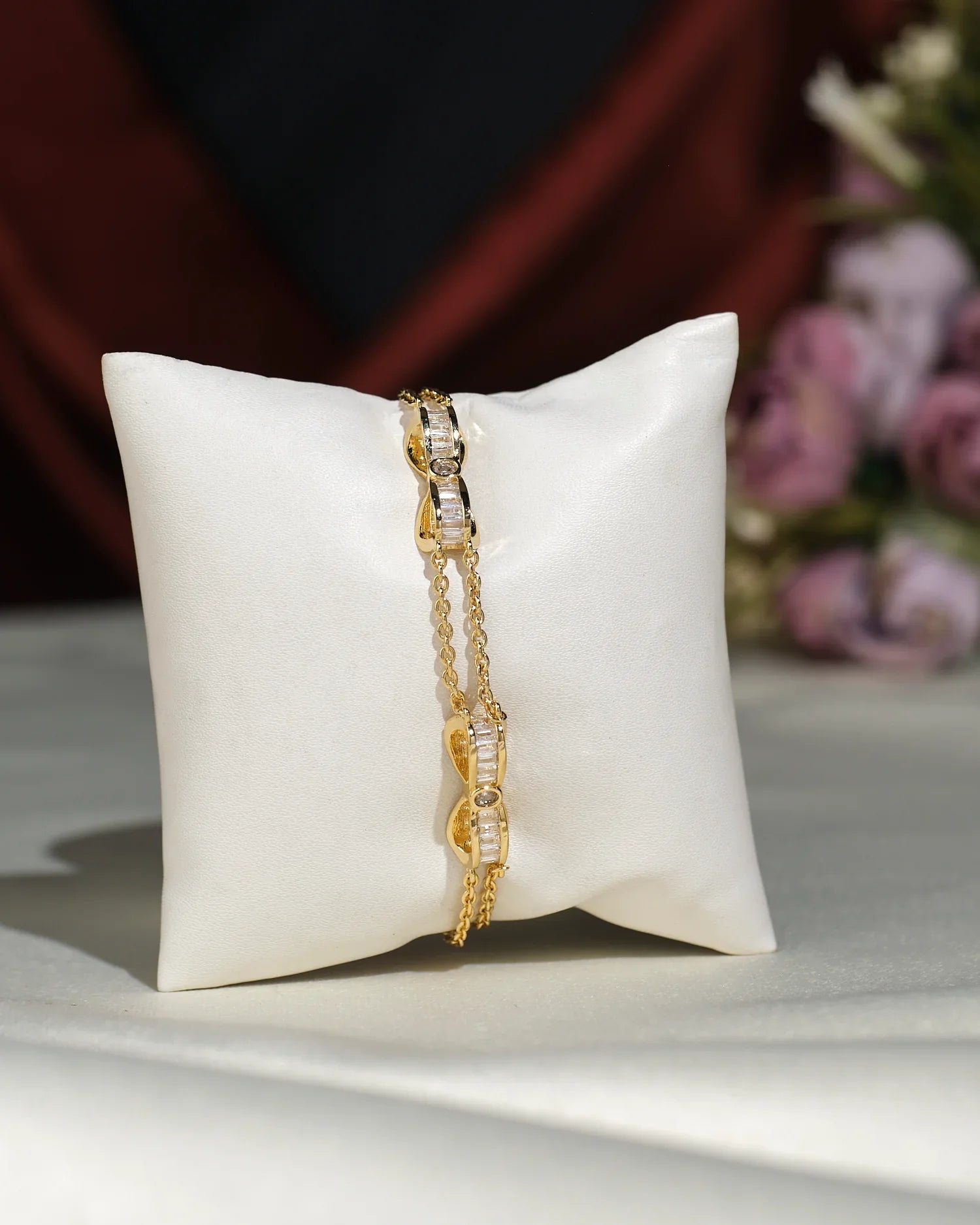 The Metallic CZ Pretty Bracelet by Chandrani Pearls India, featuring clear stone accents, rests on a white cushion with blurred purple flowers in the background, creating an elegant and charming display.