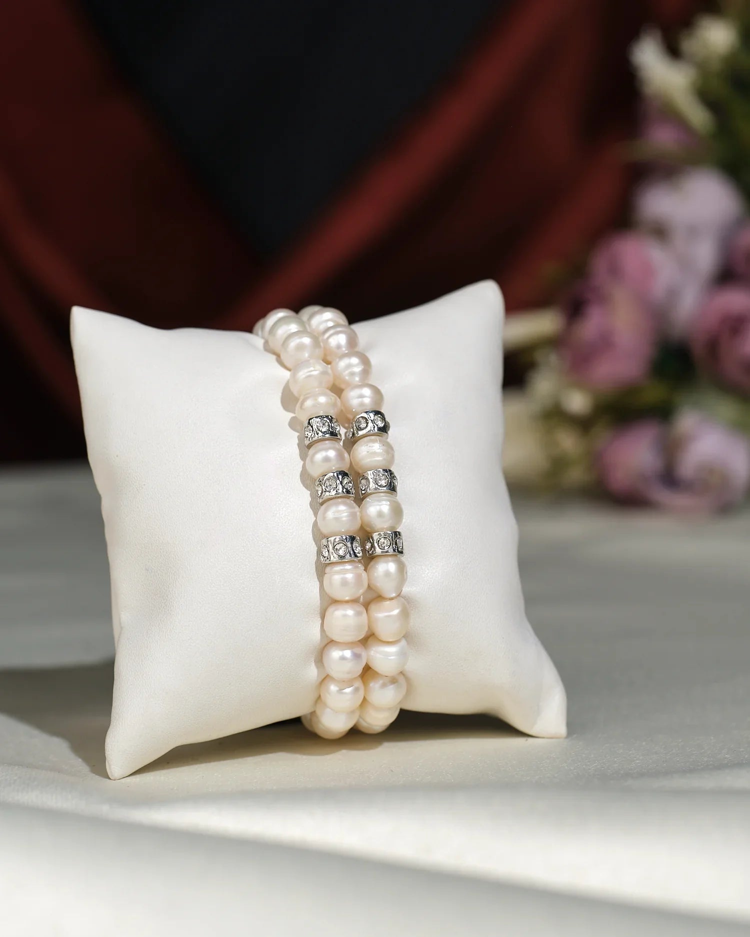 The Elegant White 2 line Bracelet by Chandrani Pearls India features double strands with pearl beads and silver spacers, displayed on a white cushion, accented by blurred flowers in the background for a unique look.