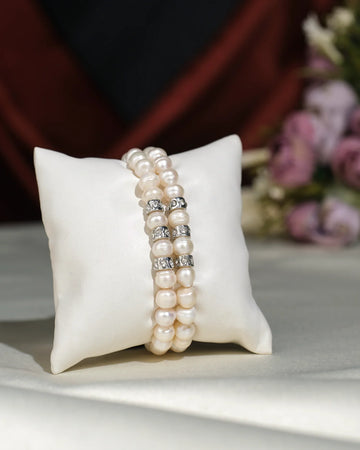 The Elegant White 2 line Bracelet by Chandrani Pearls India features double strands with pearl beads and silver spacers, displayed on a white cushion, accented by blurred flowers in the background for a unique look.