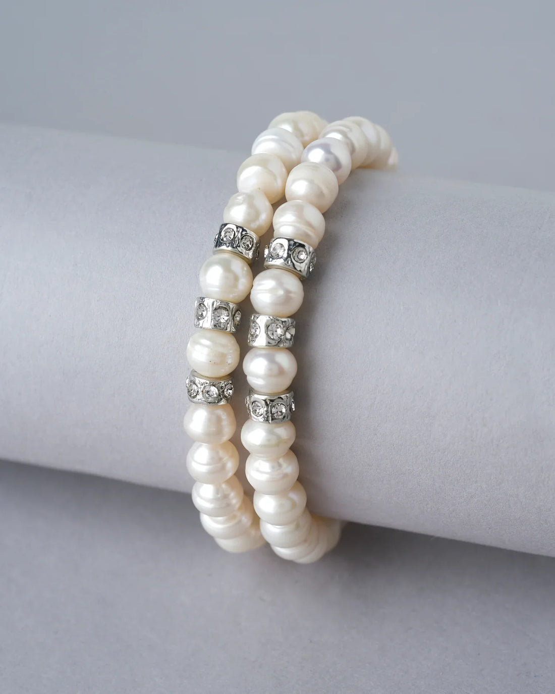 The Elegant White 2 line Bracelet by Chandrani Pearls India features double strands with pearl beads and silver spacers, displayed on a white cushion, accented by blurred flowers in the background for a unique look.