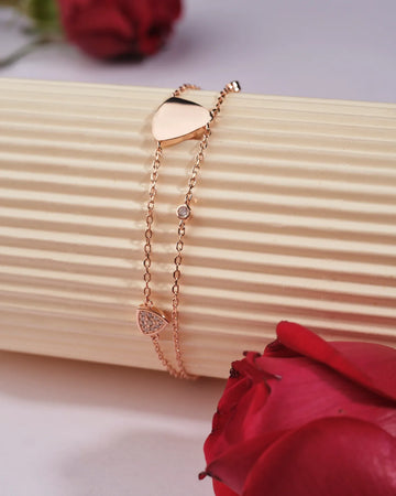 The Tamanna 925 Silver Rose Gold Bracelet from Chandrani Pearls India, featuring heart and triangle charms in a polished, unique style, is draped over a ridged cream cylinder with red roses nearby.