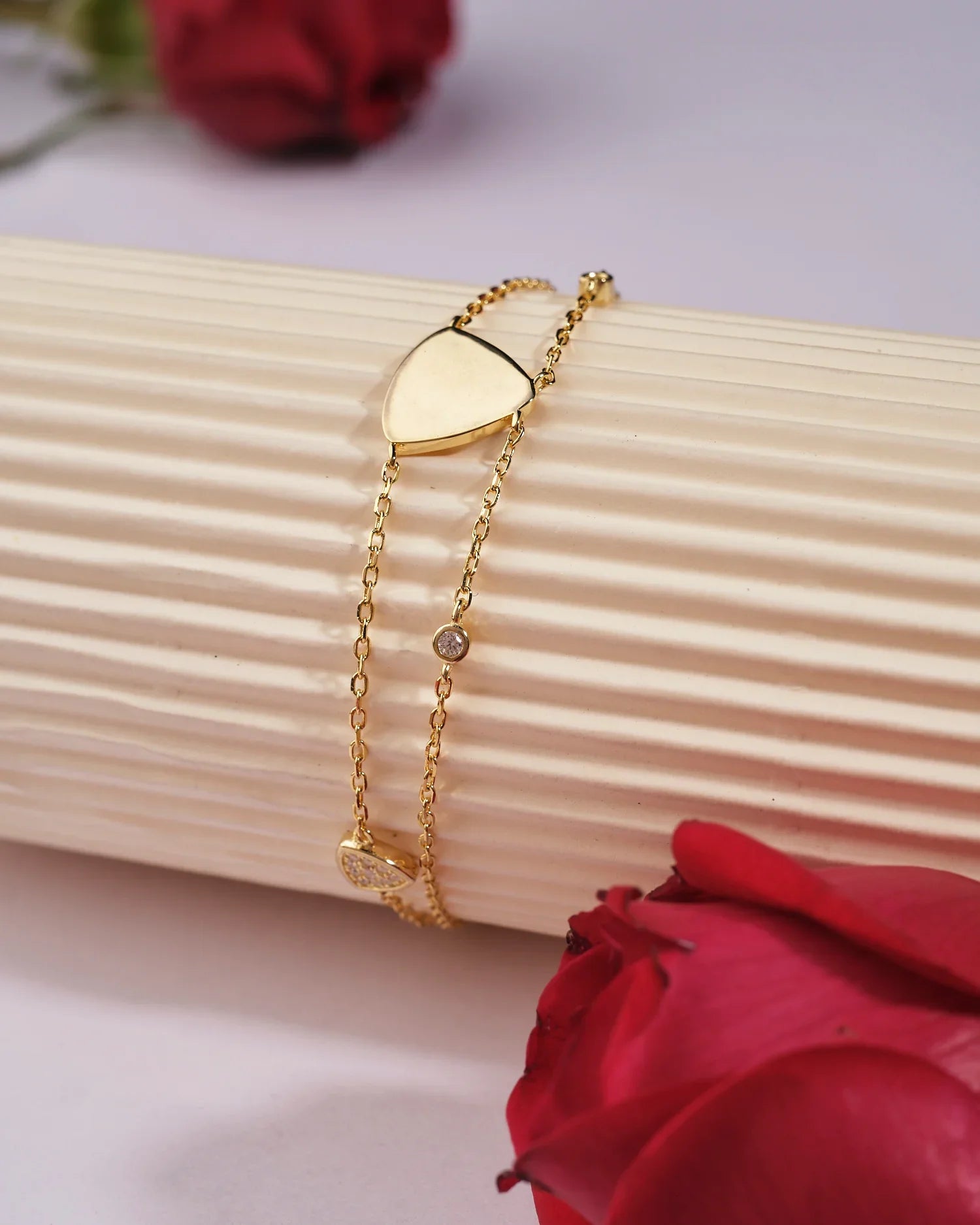 The Triya 925 Silver Gold Bracelet by Chandrani Pearls India, featuring a heart charm and round gem, adds elegance; shown on a ribbed cream surface with red roses in the background.