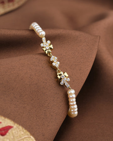The Noor Soft Glow Bracelet by Chandrani Pearls, featuring delicate pearls and floral CZ accents, is displayed on a brown fabric background.