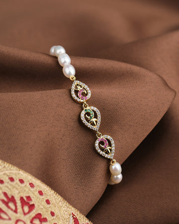 The Noor Graceful Glam Bracelet by Chandrani Pearls features heart motifs, red and green stones, and natural pearls, adding elegance to your look. Shown on brown fabric with a touch of red and gold embroidery.
