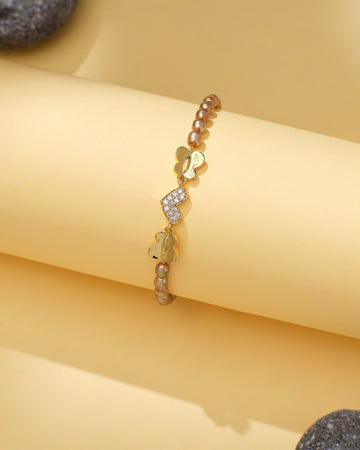 Chandrani Pearls' Elegant Pearl & CZ Tennis Bracelet in Brown Pearl features butterfly charms, some with sparkling crystals, and metallic beads, displayed on a light yellow background.