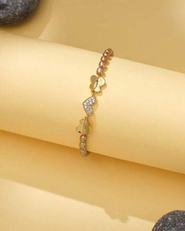 Chandrani Pearls' Elegant Pearl & CZ Tennis Bracelet in Brown Pearl features butterfly charms, some with sparkling crystals, and metallic beads, displayed on a light yellow background.