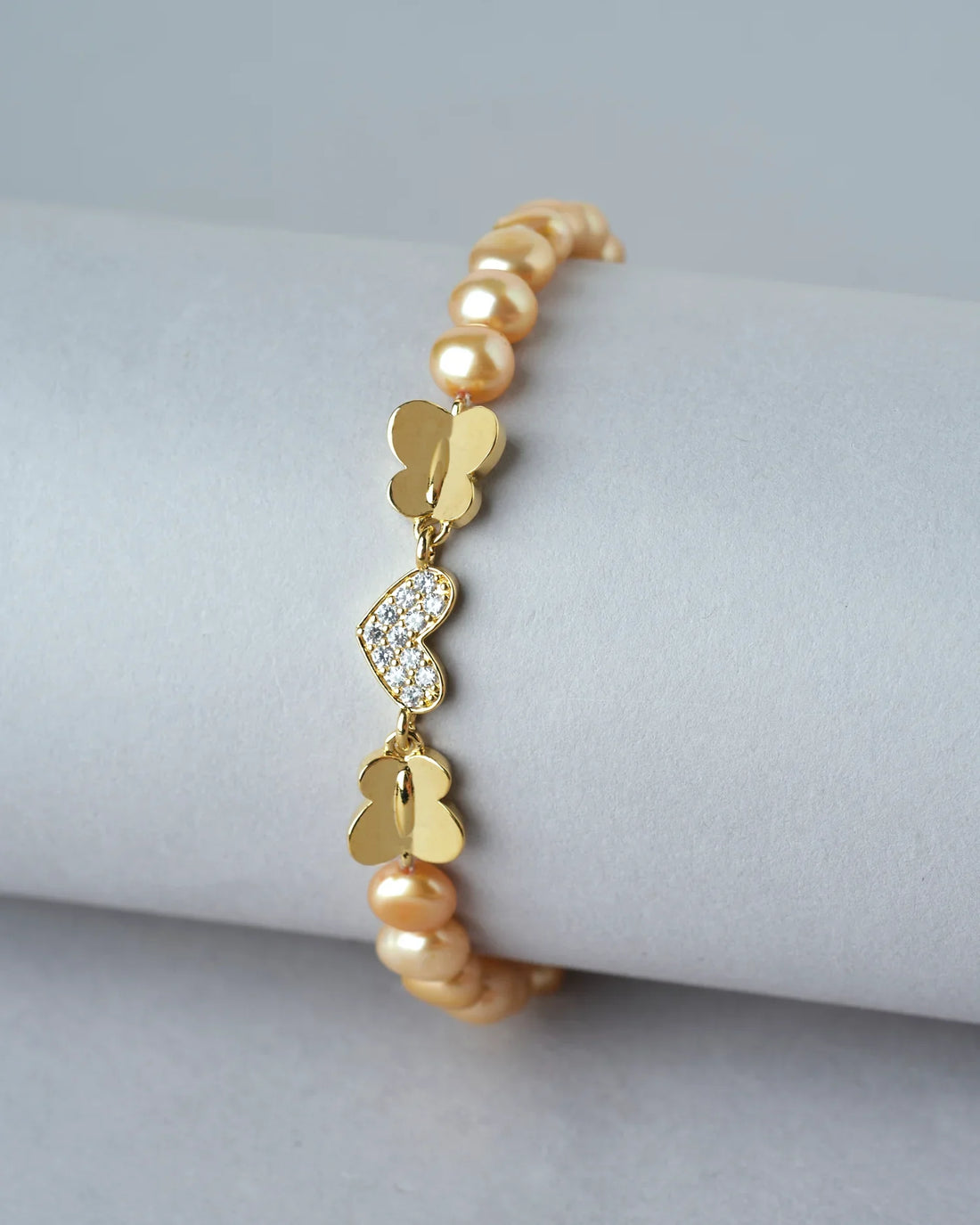 Little Heart CZ Pearl Bracelet by Chandrani Pearls India shown on a white cushion, featuring pearls and a decorative clasp, with blurred flowers in the background.