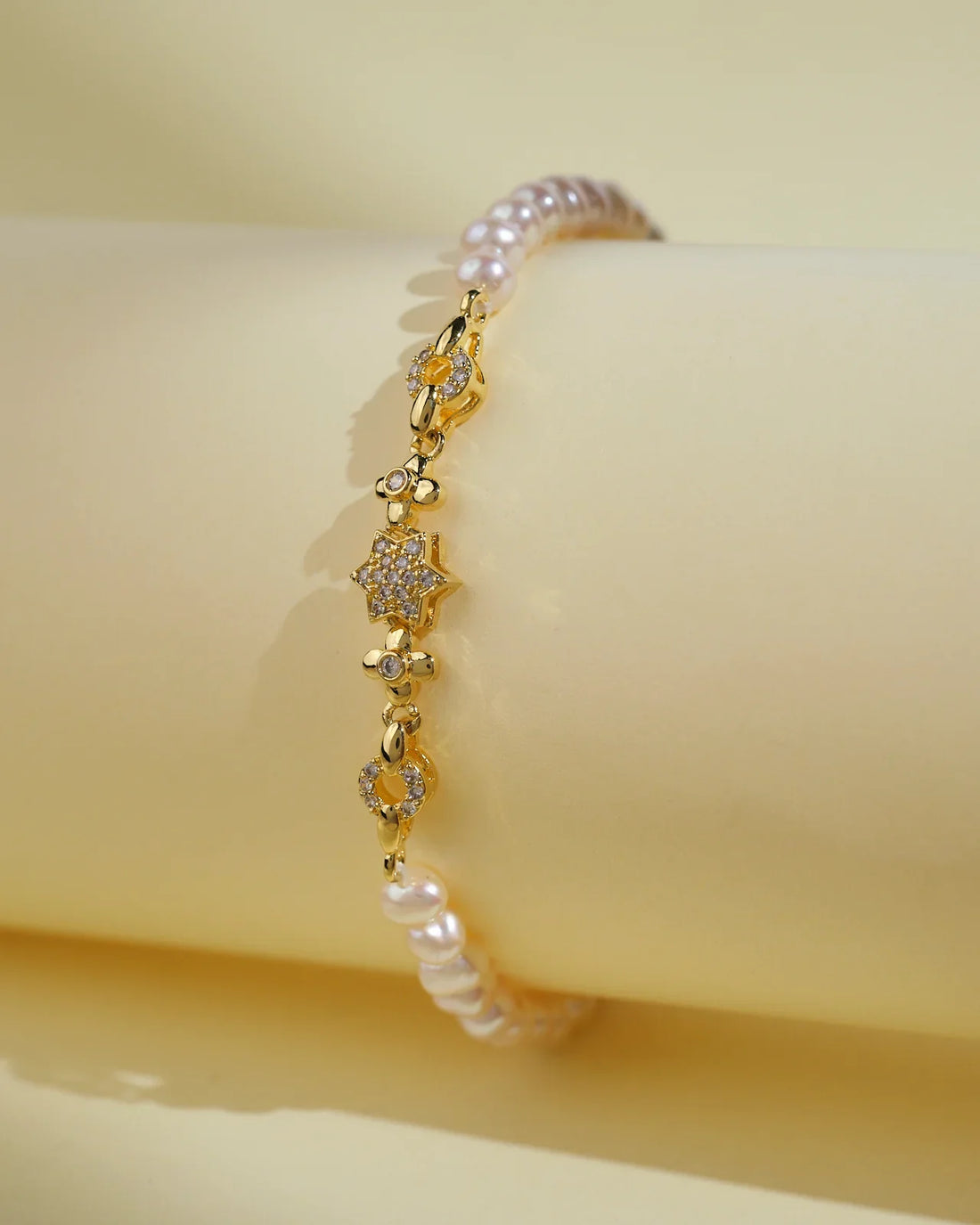 The Flory White 1 Line Pearl Bracelet by Chandrani Pearls India features gold, small floral designs, and clear stones for everyday elegance. Shown against a pale yellow background.