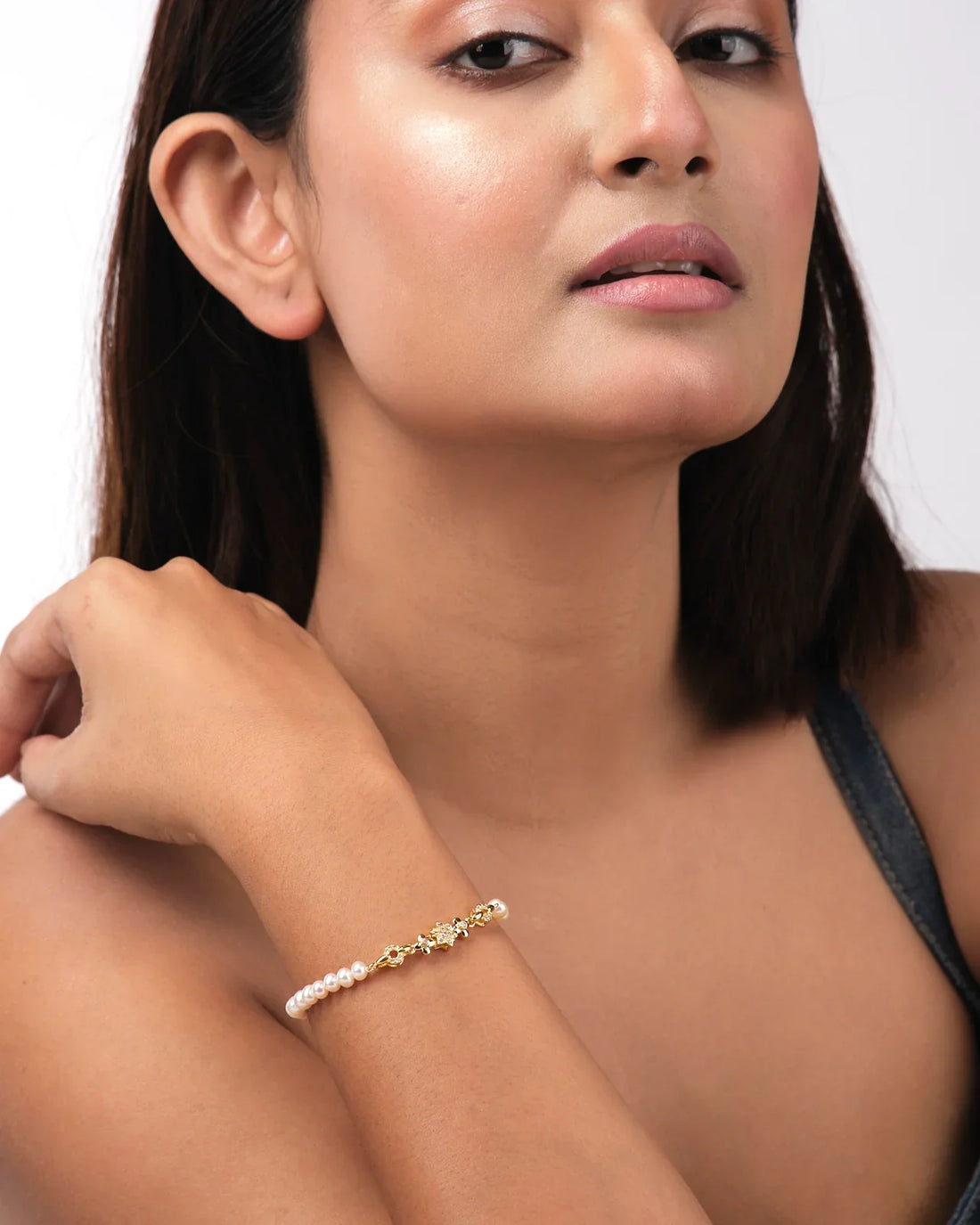 The Flory White 1 Line Pearl Bracelet by Chandrani Pearls India features gold, small floral designs, and clear stones for everyday elegance. Shown against a pale yellow background.