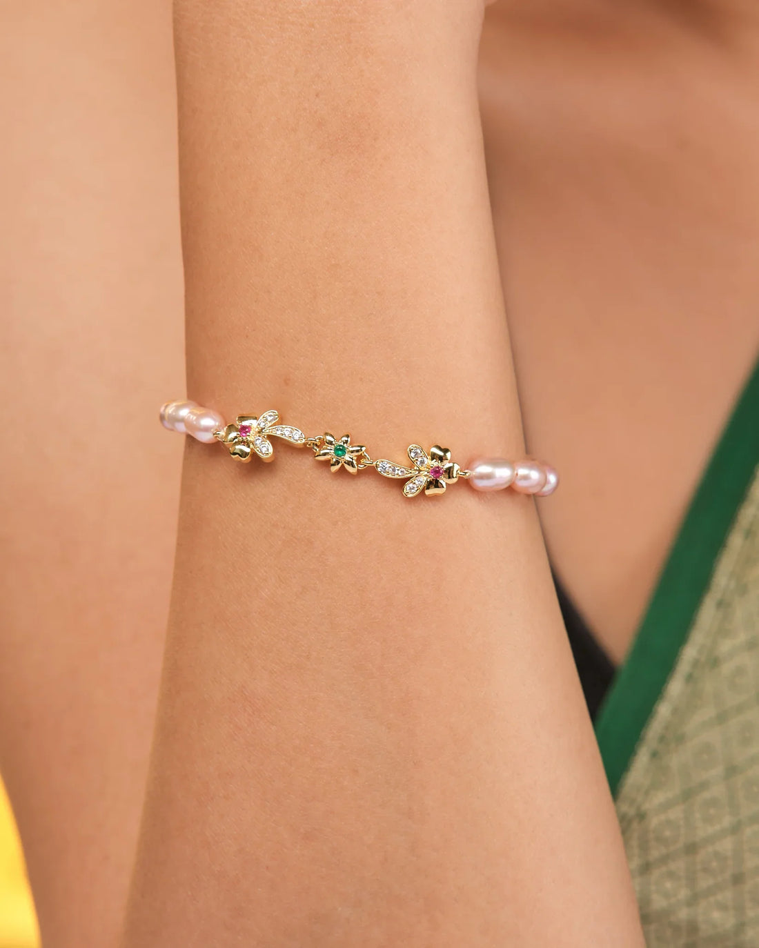 The Noor Classic Pearl Radiance Bracelet by Chandrani Pearls features hand-selected pearls, CZ stones, and gold floral and leaf charms, elegantly displayed on brown fabric with a gold embroidered edge.