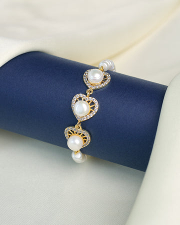 The Modern Pearl Bracelet with Cubic Zirconia Highlights from Chandrani Pearls features three gold, heart-shaped links adorned with pearls and sparkling stones—an elegant choice for wedding jewelry, shown on a dark blue display against light fabric.