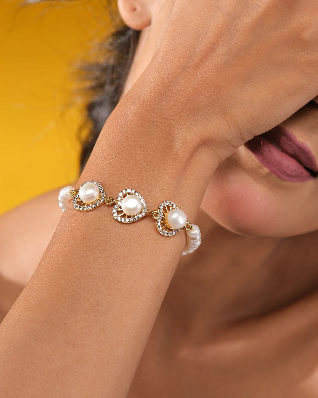The Modern Pearl Bracelet with Cubic Zirconia Highlights from Chandrani Pearls features three gold, heart-shaped links adorned with pearls and sparkling stones—an elegant choice for wedding jewelry, shown on a dark blue display against light fabric.