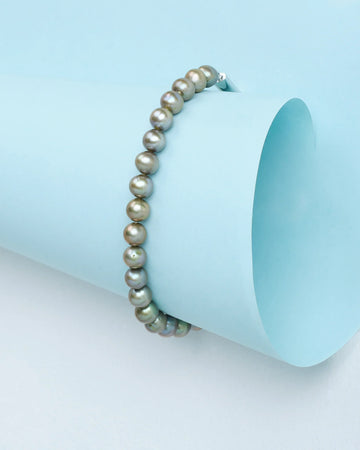 The "Pearls that speak elegance" bracelet by Chandrani Pearls, featuring round metallic-hued pearls, is displayed on light blue paper and adds chic sophistication to any look.
