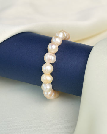 The Statement Pearl Bracelet with Dazzling CZ by Chandrani Pearls, featuring round white pearls and sparkling cubic zirconia, is elegantly displayed on a dark blue cylindrical stand against a light cream fabric background.