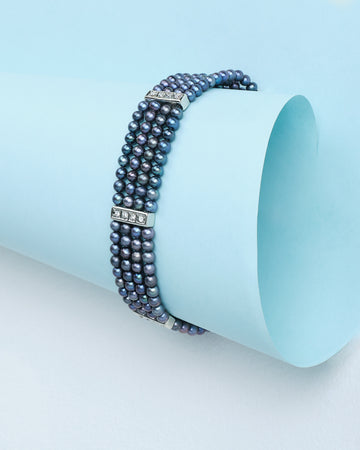 The "A Touch of Elegance on Your Wrist" bracelet by Chandrani Pearls features three rows of sophisticated pearls with rhinestone-studded spacers, displayed on a rolled sheet of light blue paper.