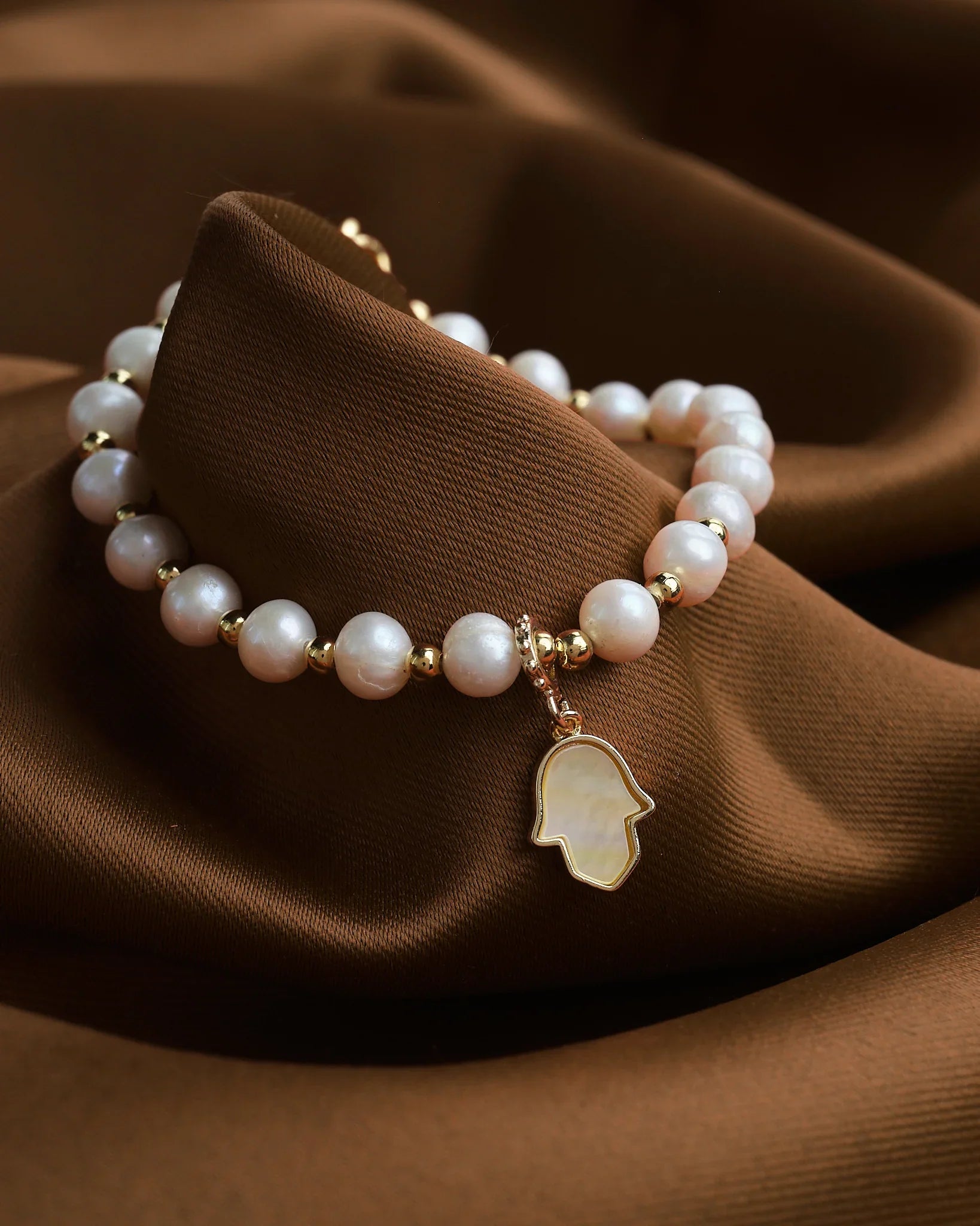 The Pretty Mother of Pearl Charm Bracelet by Chandrani Pearls features gold accents and a Hamsa hand pendant, elegantly displayed on brown fabric.