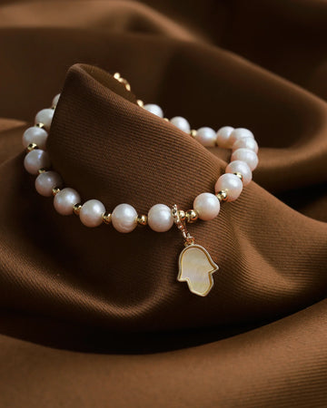 The Pretty Mother of Pearl Charm Bracelet by Chandrani Pearls features gold accents and a Hamsa hand pendant, elegantly displayed on brown fabric.