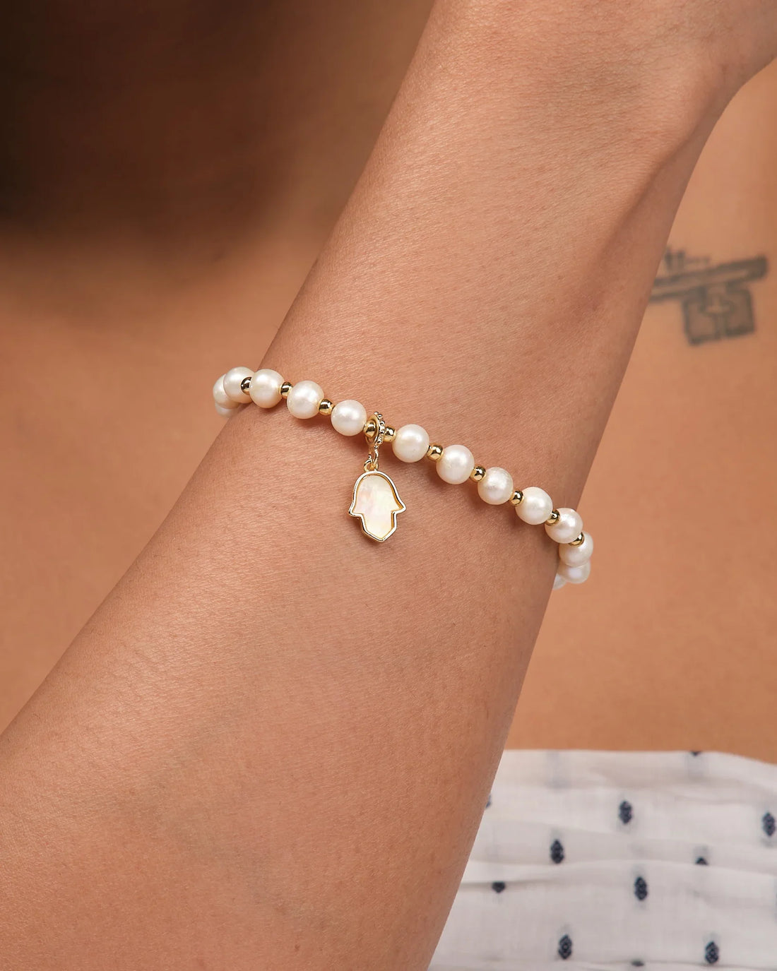 The Pretty Mother of Pearl Charm Bracelet by Chandrani Pearls features gold accents and a Hamsa hand pendant, elegantly displayed on brown fabric.