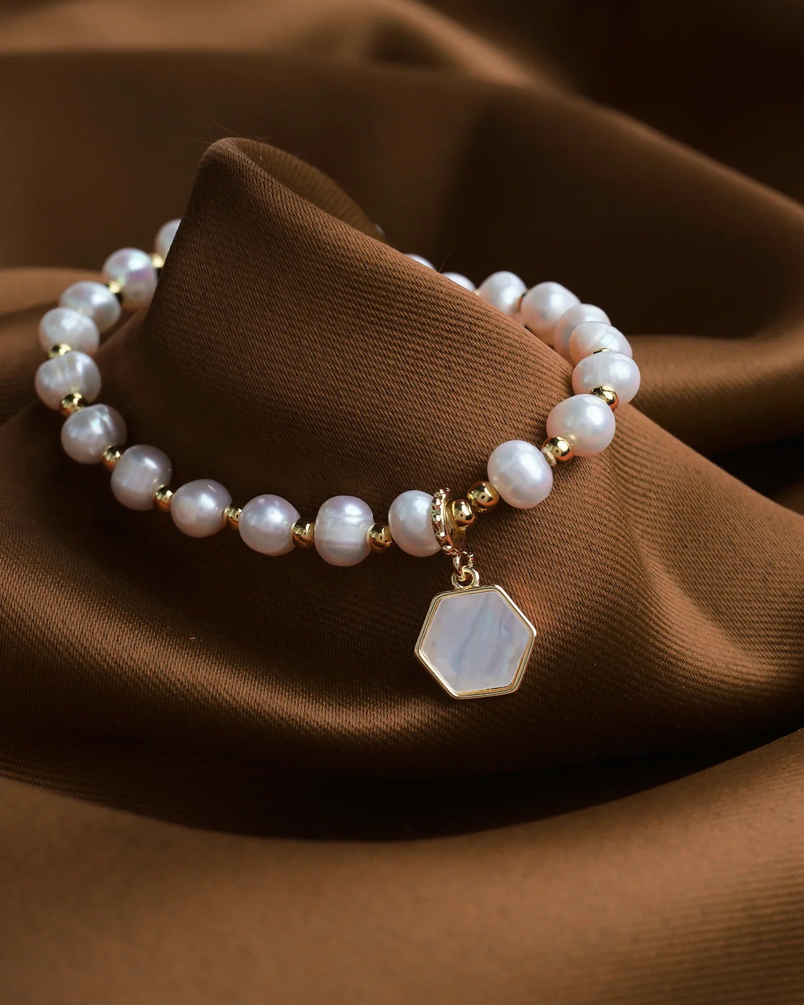 The Gleaming Touch bracelet by Chandrani Pearls, featuring pearl beads, gold accents, and a hexagonal pendant, is displayed on brown fabric—offering subtle luxury and a timeless finish to complement any outfit.