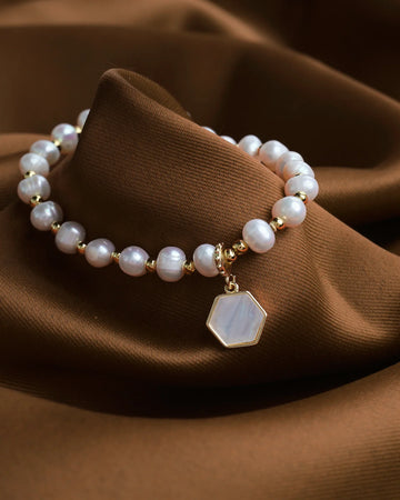 The Gleaming Touch bracelet by Chandrani Pearls, featuring pearl beads, gold accents, and a hexagonal pendant, is displayed on brown fabric—offering subtle luxury and a timeless finish to complement any outfit.
