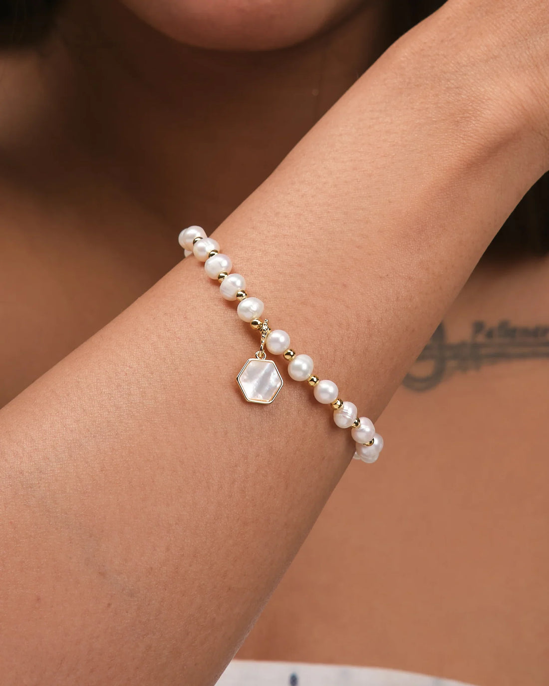 The Gleaming Touch bracelet by Chandrani Pearls, featuring pearl beads, gold accents, and a hexagonal pendant, is displayed on brown fabric—offering subtle luxury and a timeless finish to complement any outfit.