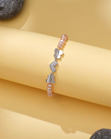 The Chandrani Pearls Elegant Pearl & CZ Tennis Bracelet Pink Pearl features pink pearls, silver butterfly charms, and sparkling cubic zirconia, displayed on a curved yellow surface with gray stones nearby.