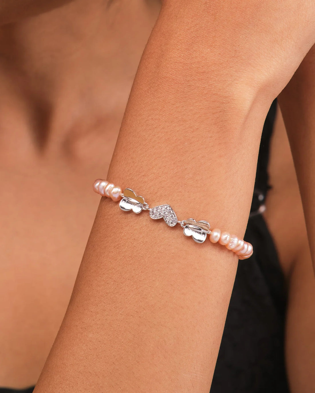 The Chandrani Pearls Elegant Pearl & CZ Tennis Bracelet Pink Pearl features pink pearls, silver butterfly charms, and sparkling cubic zirconia, displayed on a curved yellow surface with gray stones nearby.