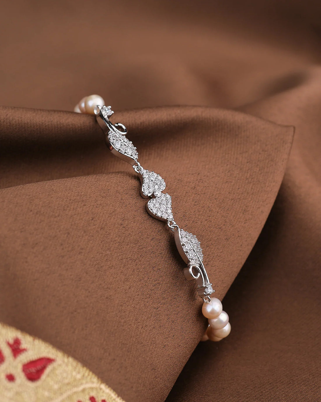 The Noor Leafy CZ Pearl Bracelet by Chandrani Pearls features heart and leaf designs with sparkling CZ accents and delicate pearls, shown on brown fabric.