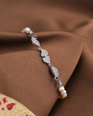 The Noor Leafy CZ Pearl Bracelet by Chandrani Pearls features heart and leaf designs with sparkling CZ accents and delicate pearls, shown on brown fabric.