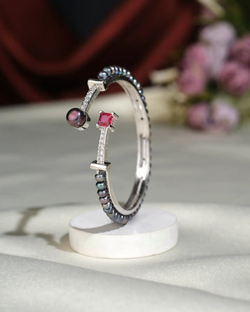 The Trendy Black Pearl Bangle by Chandrani Pearls India, featuring a silver band with a red square gemstone, round glowing pearl, black beads, and clear stones, is displayed upright on a white circular stand.