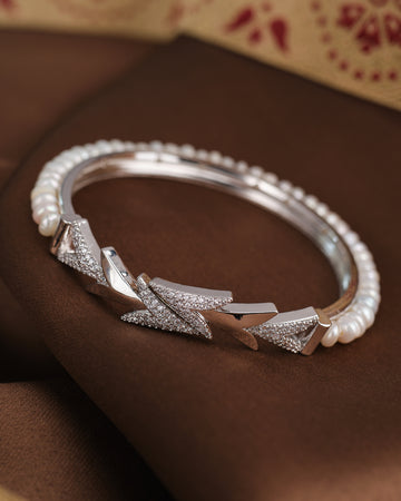 The Pearl Radiance CZ Bangle by Chandrani Pearls features geometric patterns, embedded CZ stones, and pearl accents, displayed on a brown fabric background.