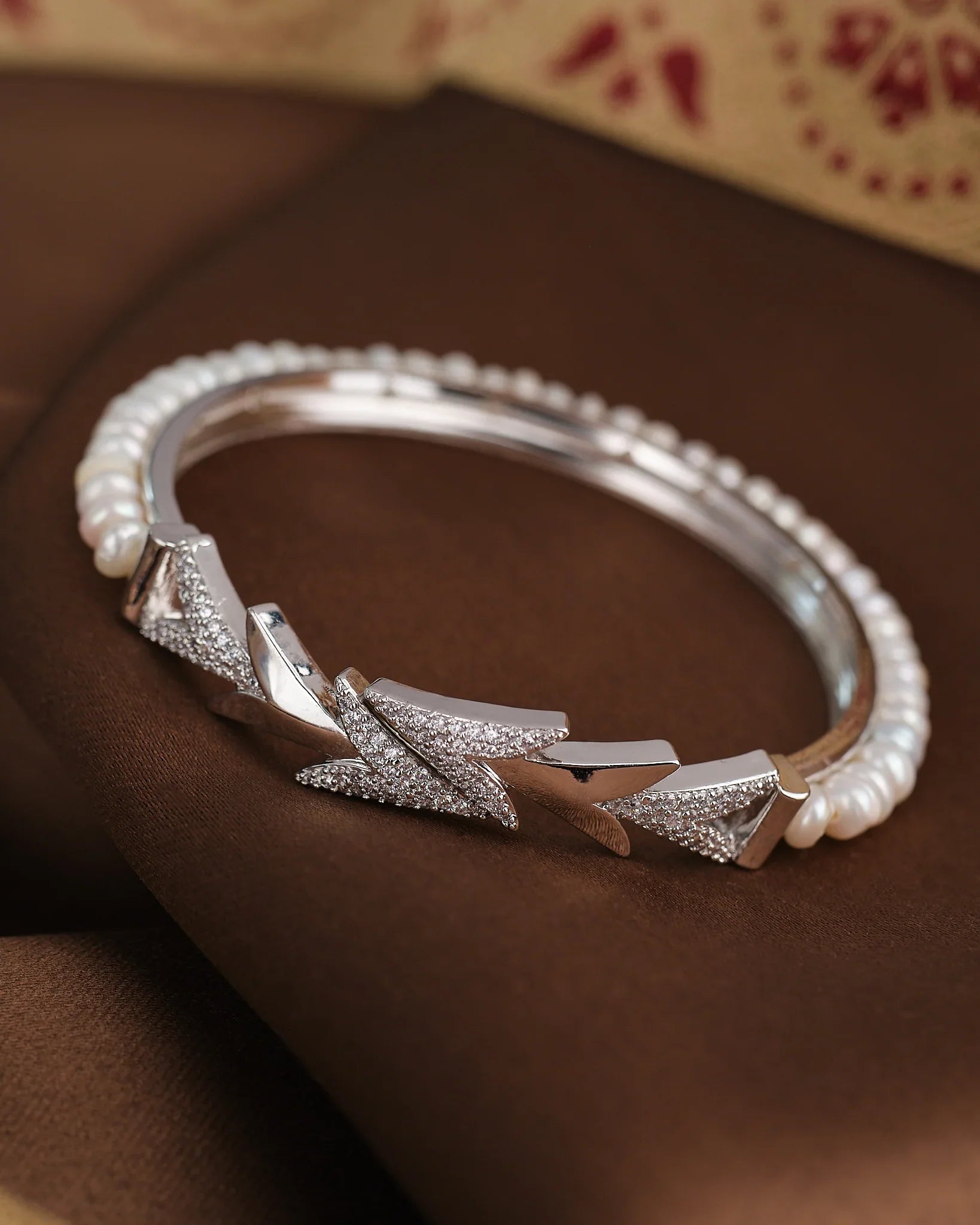 The Pearl Radiance CZ Bangle by Chandrani Pearls features geometric patterns, embedded CZ stones, and pearl accents, displayed on a brown fabric background.