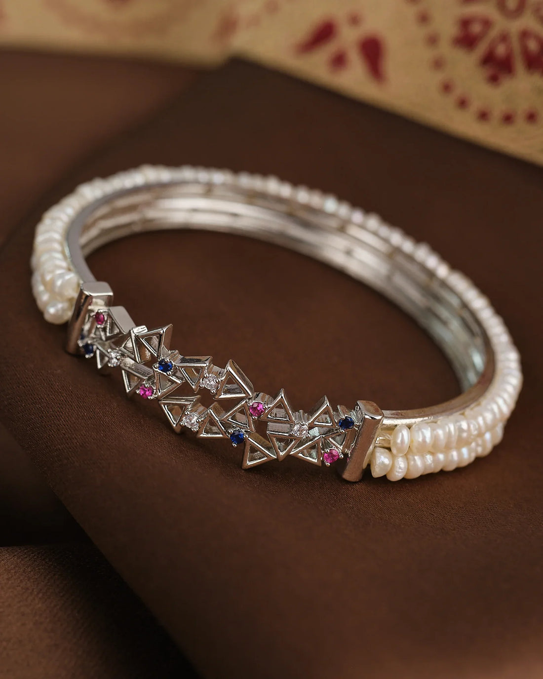 The Royal Pearl Sparkle Bangle by Chandrani Pearls features geometric silver shapes, sparkling CZ stones, and a row of white pearls, beautifully displayed on a brown fabric background.