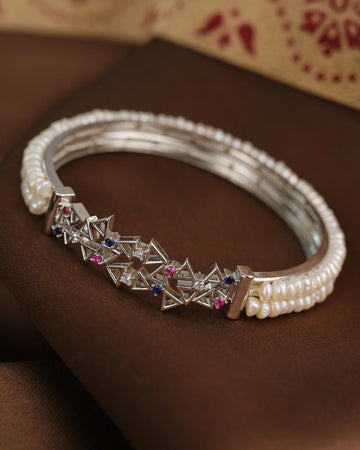 The Royal Pearl Sparkle Bangle by Chandrani Pearls features geometric silver shapes, sparkling CZ stones, and a row of white pearls, beautifully displayed on a brown fabric background.