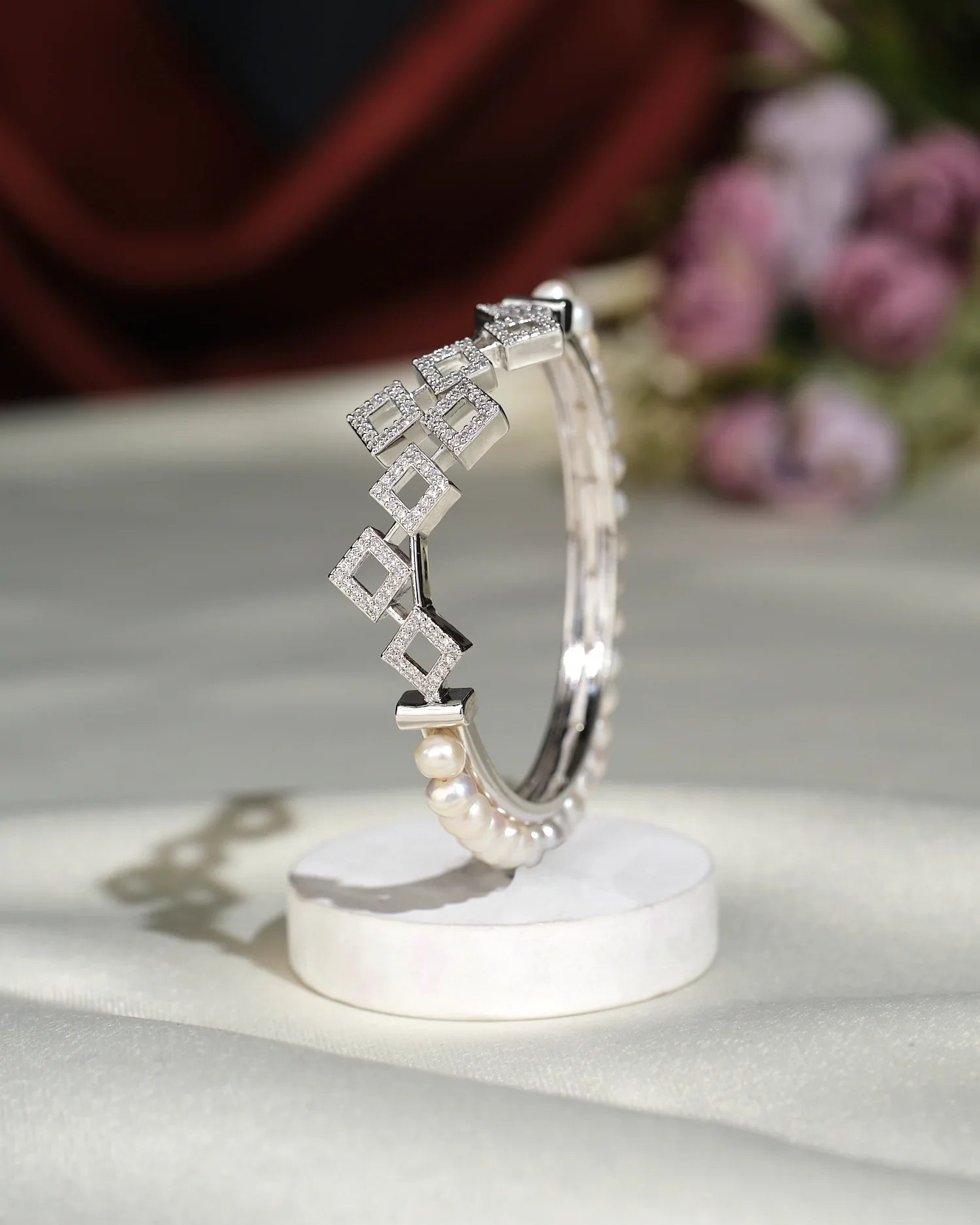 The Beautiful White Pearl Bangle by Chandrani Pearls India, featuring shiny geometric square designs and elegant pearls, is displayed upright on a round white stand with blurred flowers in the background.