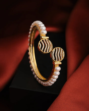 The Beautiful and Classy Pearl Bangle by Chandrani Pearls features lustrous pearls and diamond-studded wavy ends, displayed on a black surface with a red fabric background—perfect to elevate your style.