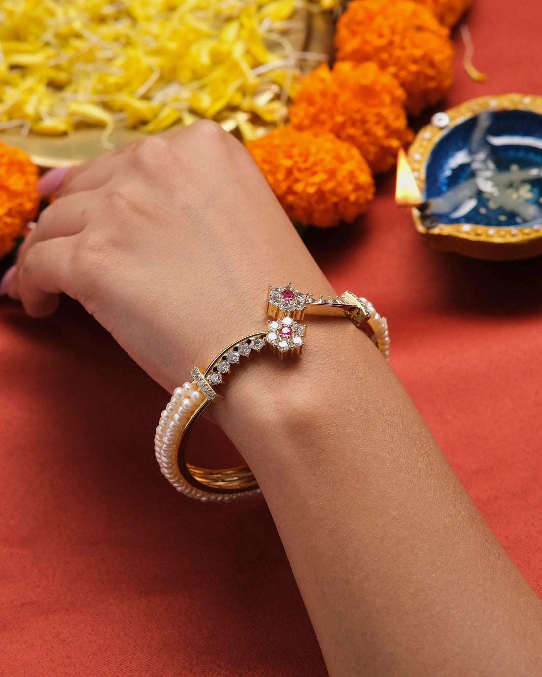 The Ragapushpi - The Rosy Night Pearl Bangle by Chandrani Pearls, adorned with pearl accents and floral embellishments, featuring pink and clear stones, is elegantly displayed on a red box against a blurred background.
