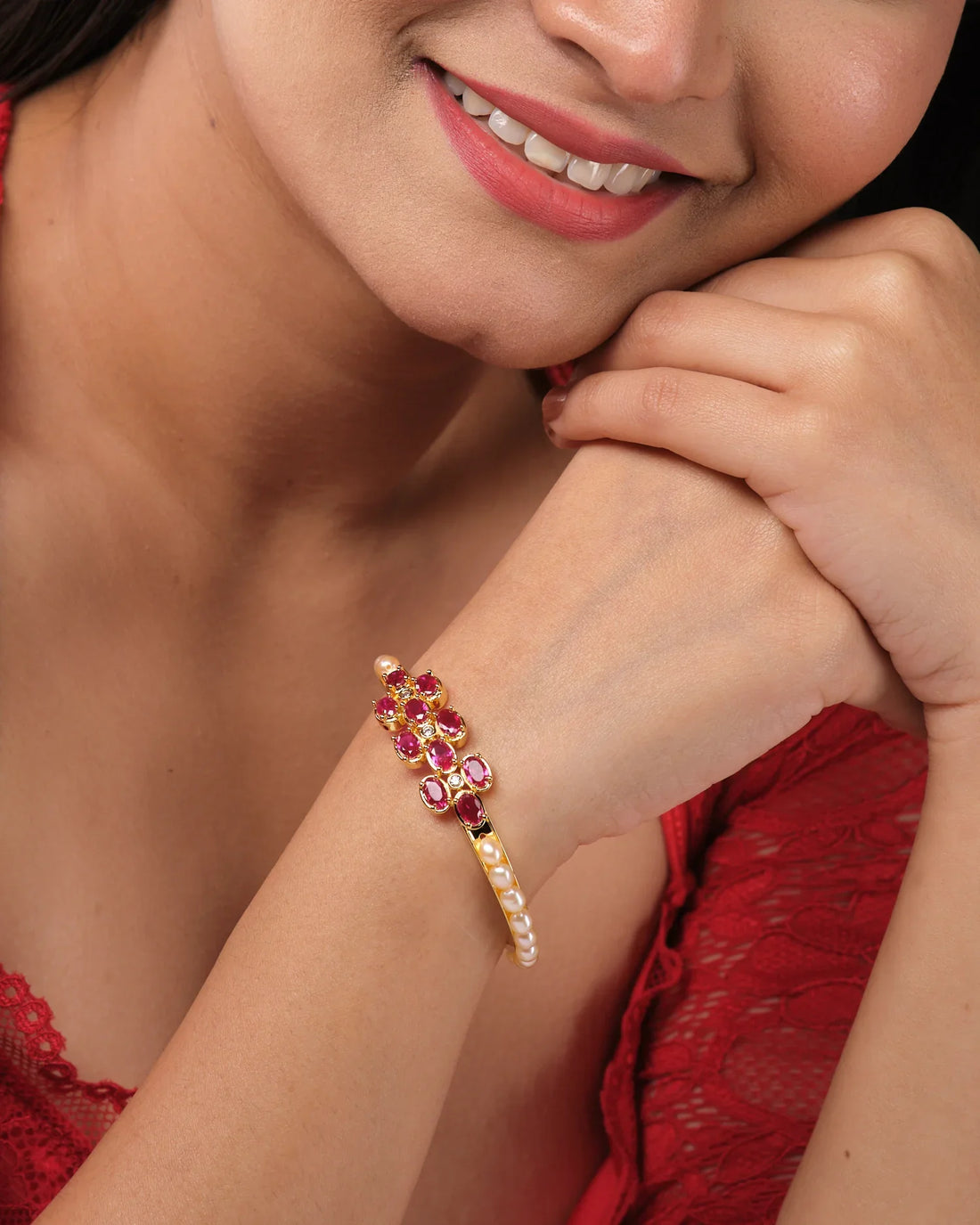 The Pretty Pink Bangle by Chandrani Pearls India, featuring pink gemstones and white pearls set in elegant gold, stands upright on a round white base against a blurred floral background.