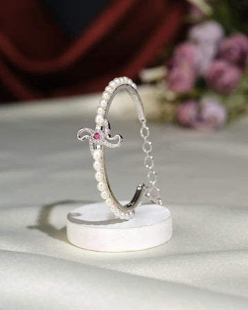 The Trendy White Bangle by Chandrani Pearls India features an elegant silver design with a pink gemstone centerpiece, displayed on a white stand with blurred flowers in the background.