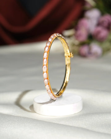 The Simple Pearl Bangle by Chandrani Pearls India, featuring inset pearls, is displayed upright on a white circular stand with a softly blurred floral background.