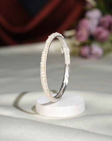 The Simple Pearl Bangle by Chandrani Pearls India features a silver design with pearls, elegantly displayed upright on a white stand against a soft purple floral backdrop—an ideal accessory to elevate your style.