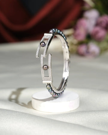 The Beautiful Black Pearl Bangle by Chandrani Pearls India features black pearls and diamond accents in a chic silver design, shown upright on a white circular stand with blurred flowers adding an elegant touch to its sophisticated appeal.